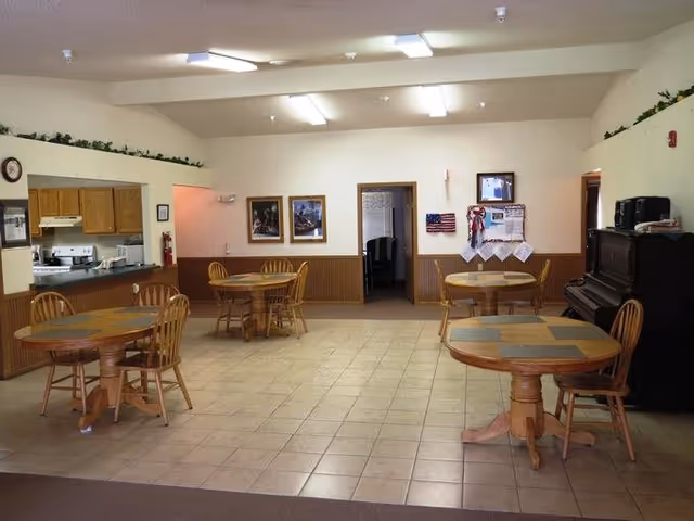A communal dining area with four round wooden tables, each surrounded by wooden chairs. The room has tiled flooring and beige walls with wood paneling halfway up. On the left side, there is a kitchen pass-through window with a stove and cabinets visible. The back wall has two framed pictures, a bulletin board with decorations, and an American flag. On the right side, there is a black upright piano with a few items on top. The ceiling has fluorescent lighting and some greenery decor along the top of the walls.