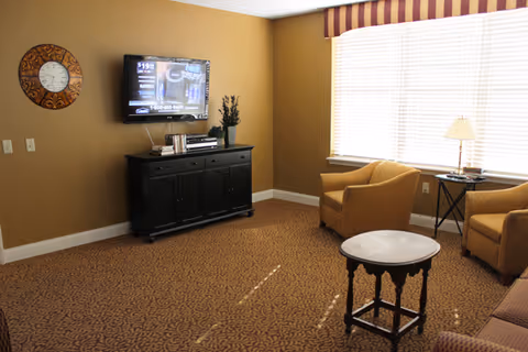 Living room with a wall-mounted TV above a black cabinet, two upholstered armchairs by a large window, and a small round coffee table.