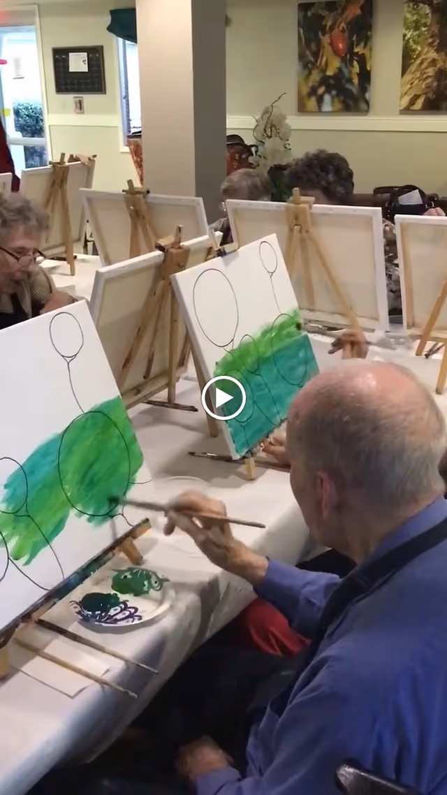 A group of elderly individuals seated at a long table in a room, each painting on canvases set on easels. The paintings show outlines of balloons and a green landscape being filled in with paint. The setting appears to be a communal activity space with artwork on the walls and a window in the background.