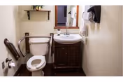Small bathroom with a white toilet equipped with a grab bar on the left side, a white sink with a dark wooden cabinet underneath, a mirror above the sink, a paper towel dispenser on the right wall, and a shelf with a small plant above the toilet.