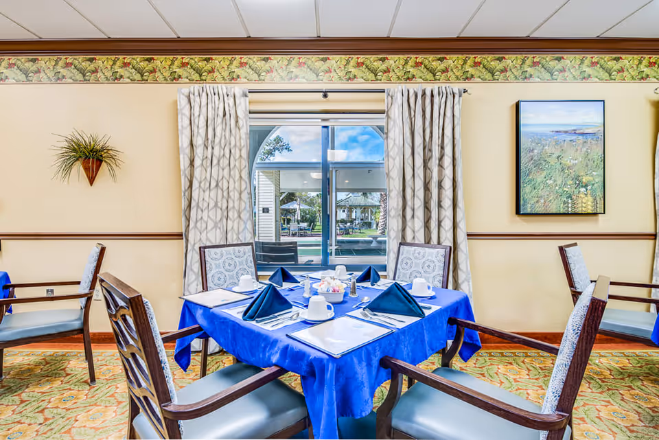 A dining room with a square table covered with a blue tablecloth, set with white cups, plates, silverware, and blue folded napkins. Four wooden chairs with cushioned seats surround the table. The room has patterned curtains framing a window that looks out onto an outdoor patio area. The walls are decorated with a floral border near the ceiling, a wall-mounted plant, and a framed landscape painting.