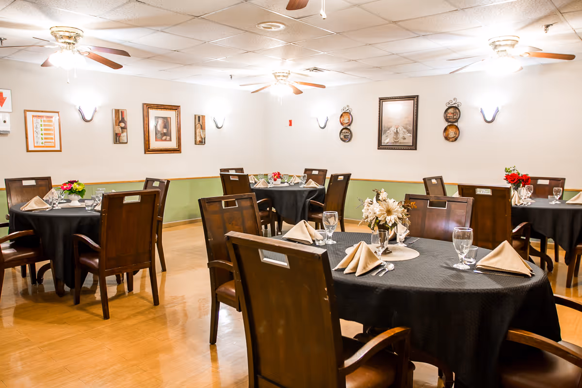 A dining room with round tables covered in black tablecloths, each set with beige folded napkins, glassware, and floral centerpieces. The room has wooden chairs, light wood flooring, ceiling fans with lights, and framed artwork on the walls.
