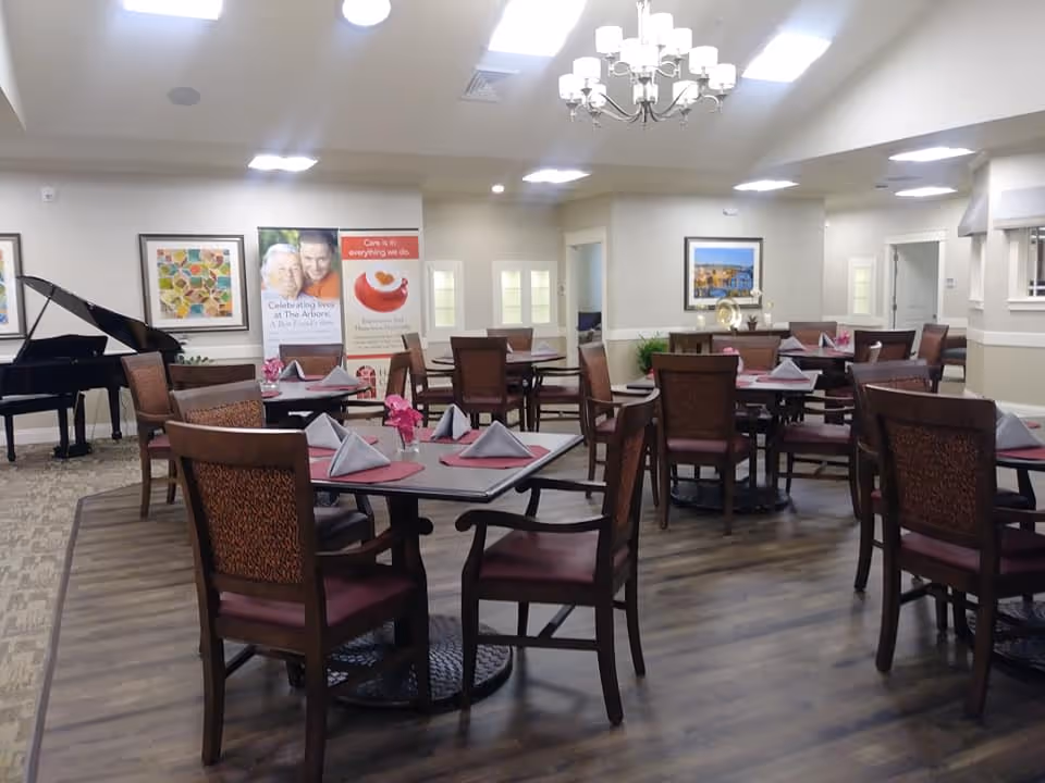 A dining room in a senior living facility with several square tables set with folded napkins and small flower centerpieces. The room has wooden chairs with cushioned seats, a grand piano in the corner, framed artwork on the walls, and a chandelier hanging from the ceiling. The floor is a combination of carpet and wood.