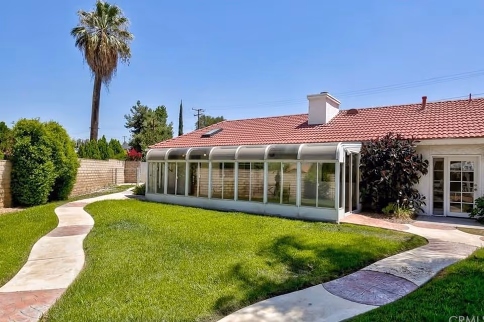 Backyard lawn with a curved walkway leading to a single-story building featuring a glass-enclosed sunroom and a red tile roof with a palm tree in the background.