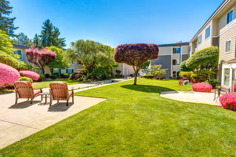 A bright and sunny outdoor courtyard area at a senior living facility with well-maintained green grass, colorful bushes, and uniquely trimmed trees. Two cushioned chairs and a small table are placed on a concrete patio, surrounded by multi-story residential buildings.