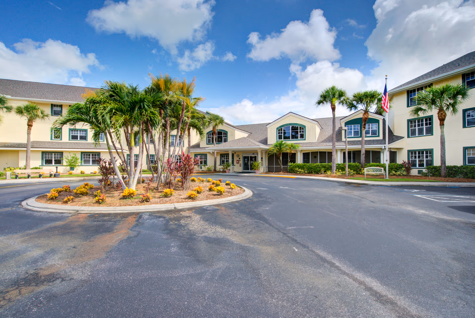 Front exterior view of Truewood by Merrill, Charlotte Center, showing a large three-story building with multiple windows, palm trees, and a circular driveway with landscaped plants in the center under a partly cloudy blue sky.