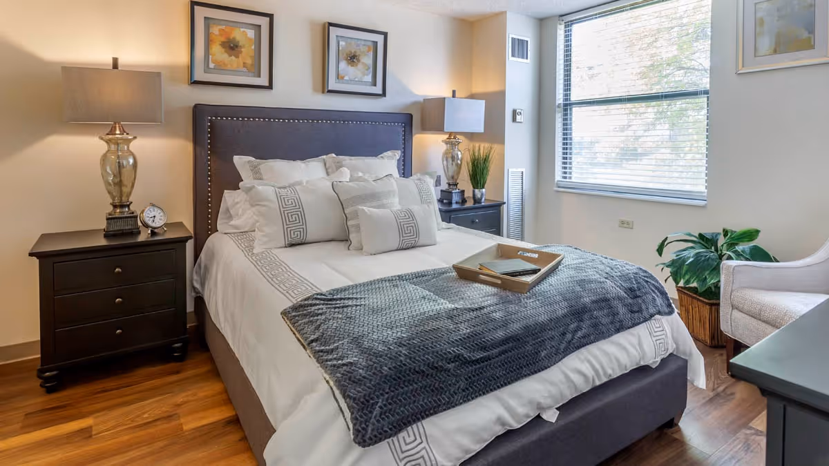 A neatly made bed with white and gray bedding, decorative pillows, and a dark gray upholstered headboard in a well-lit bedroom. There are two matching bedside tables with lamps, a small clock on the left table, framed floral artwork above the bed, a window with blinds on the right wall, a potted plant near the window, and a light-colored armchair partially visible on the right side.