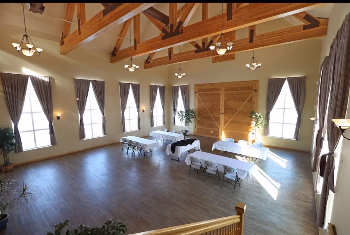 Spacious high-ceilinged event room with exposed wooden beams, tall curtained windows, and folding tables and chairs arranged on a wood floor.