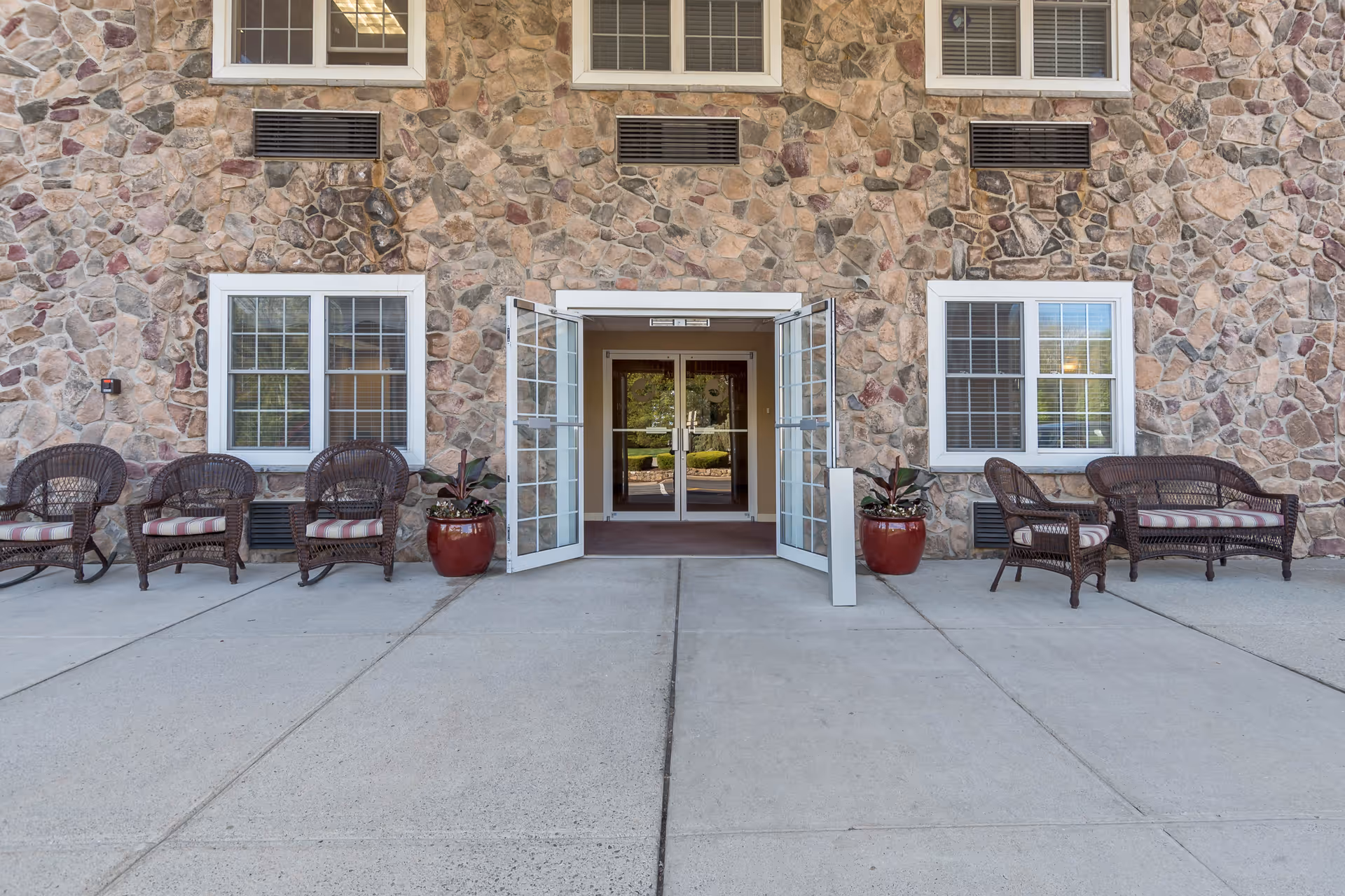 Exterior view of a building with a stone facade featuring two windows on each side of a double glass door entrance. The entrance doors are open, and there are wicker chairs and a wicker loveseat with cushions arranged on the concrete patio on both sides of the door. Two large red planters with plants are placed near the entrance.