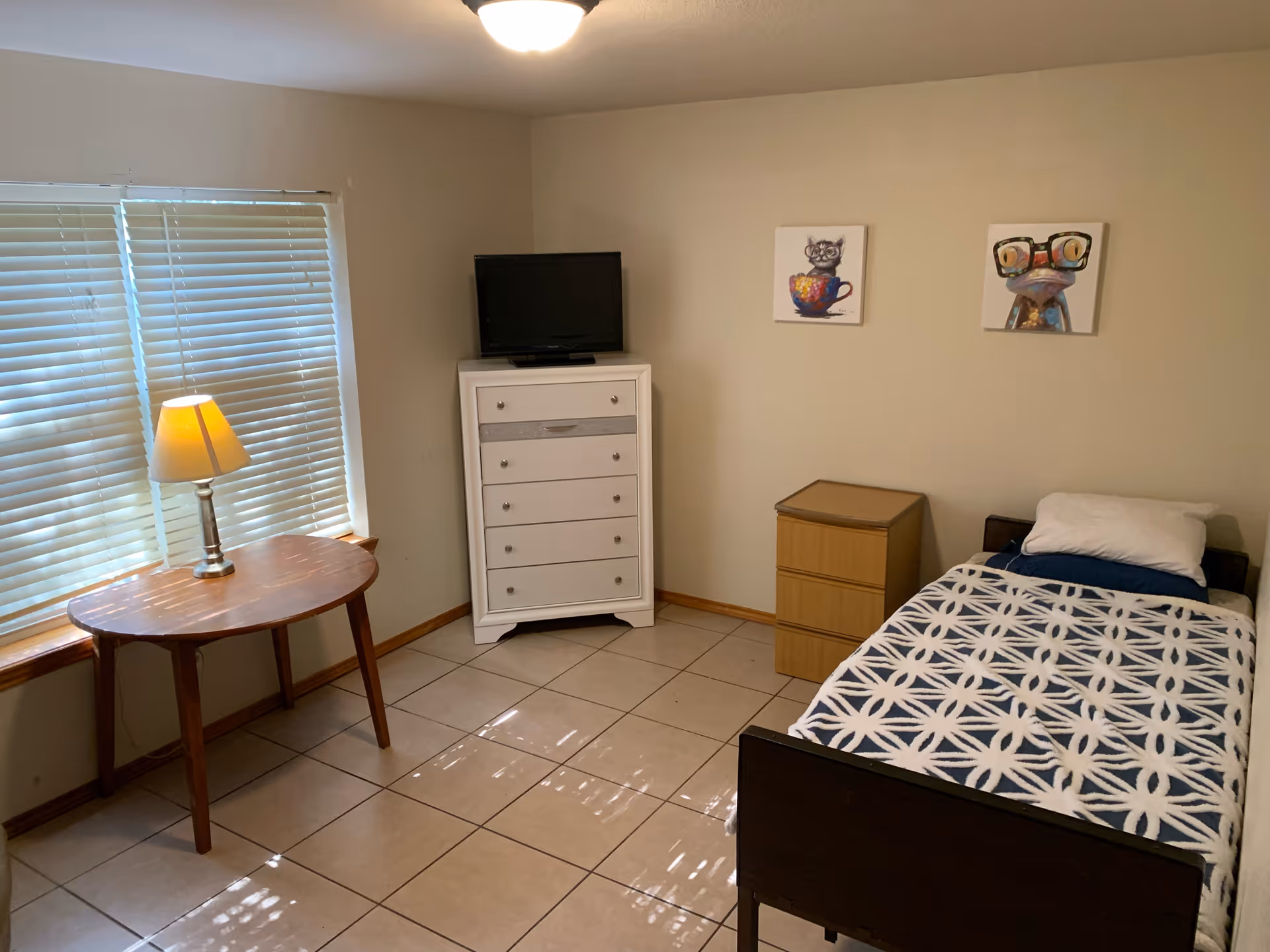 A small bedroom with a single bed covered with a blue and white patterned blanket and a white pillow. Next to the bed is a small wooden nightstand. On the wall above the nightstand are two framed pictures, one of a cat in a teacup and the other of a frog wearing glasses. Across from the bed is a white dresser with a small flat-screen TV on top. A wooden table with a lamp is placed near a window with closed blinds. The floor is tiled and the walls are painted beige.