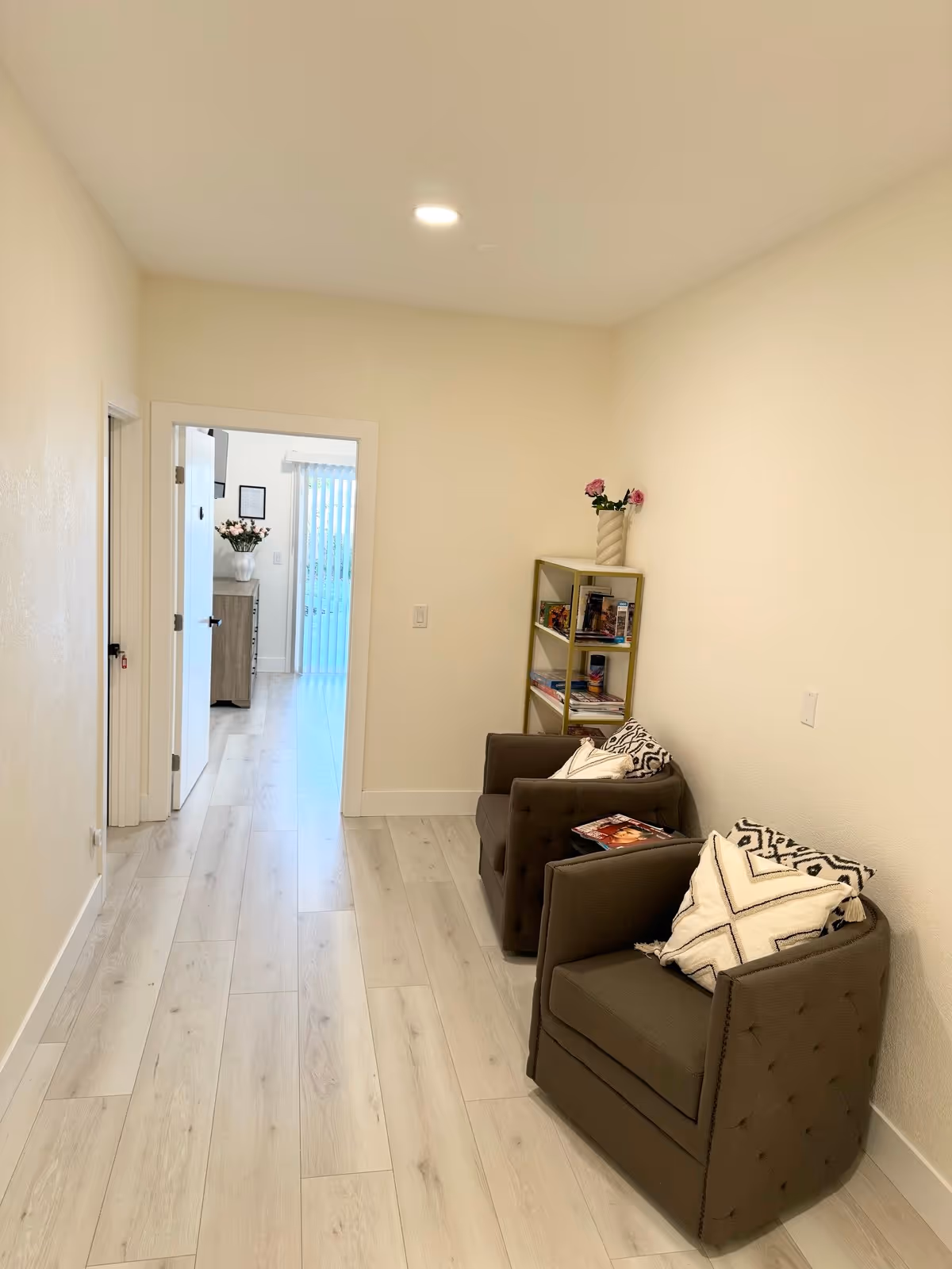 A narrow hallway with light wood flooring and cream-colored walls. Two brown armchairs with patterned cushions are placed along the right wall. A small shelving unit with books and a vase with pink flowers is positioned between the chairs. At the end of the hallway, an open door reveals another room with a dresser, a vase with flowers, and vertical blinds covering a window or door.