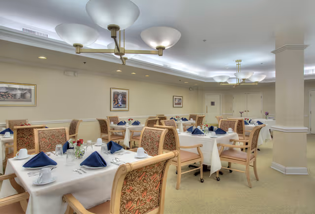 A dining room in a senior living facility with multiple tables covered in white tablecloths, each set with blue folded napkins, white cups, glasses, and silverware. The chairs have wooden frames with patterned upholstery. The room has soft lighting from ceiling fixtures and framed artwork on the walls.