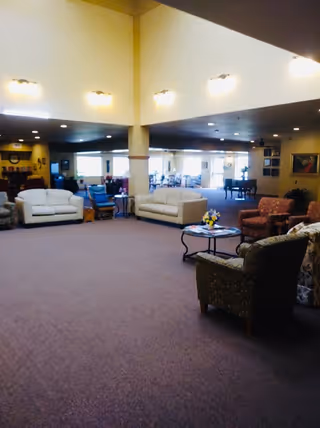 A spacious senior living facility common area with multiple seating arrangements including white sofas, patterned armchairs, and a round coffee table with a flower arrangement. The room has high ceilings with wall-mounted lights and large windows letting in natural light. In the background, there are more chairs and tables, and a piano is visible near the far wall.