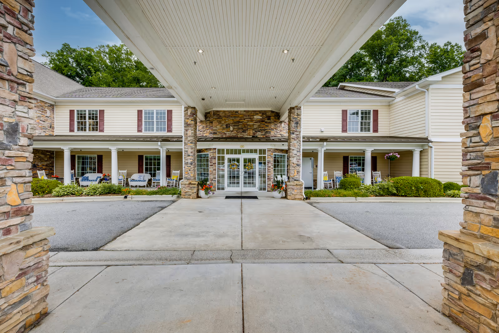 Front entrance of Arbor Ridge At Kernersville senior living facility with a covered driveway, stone pillars, and a two-story building featuring beige siding, red shutters, and a porch with rocking chairs and plants.