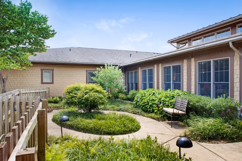 Outdoor garden area at Sunrise of Jackson featuring a curved concrete pathway surrounded by green shrubs, plants, and a tree. There is a wooden fence on the left and a metal bench with a cushion on the right near the building with multiple windows.