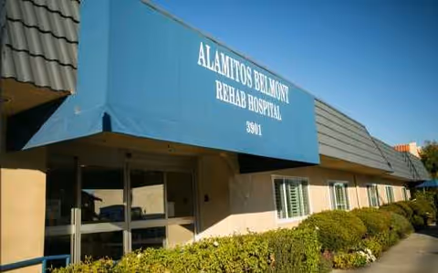 Exterior view of Alamitos Belmont Rehab Hospital entrance with blue awning and shrubs.