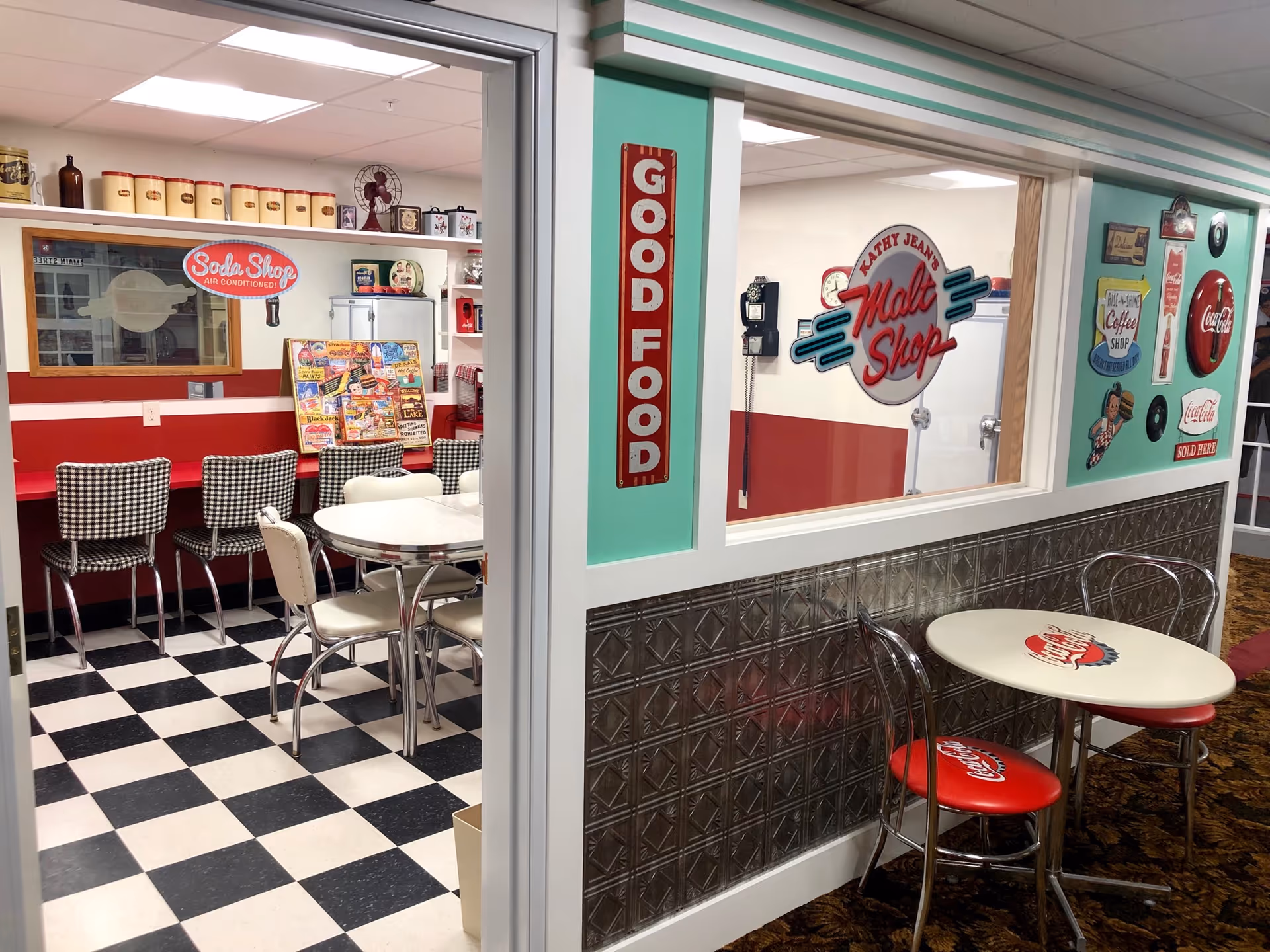 Interior view of a retro-style dining area in a senior living facility, featuring black and white checkered flooring, red and white walls, vintage soda shop signs, a counter with checkered chairs, and small round tables with Coca-Cola themed chairs and tabletops.