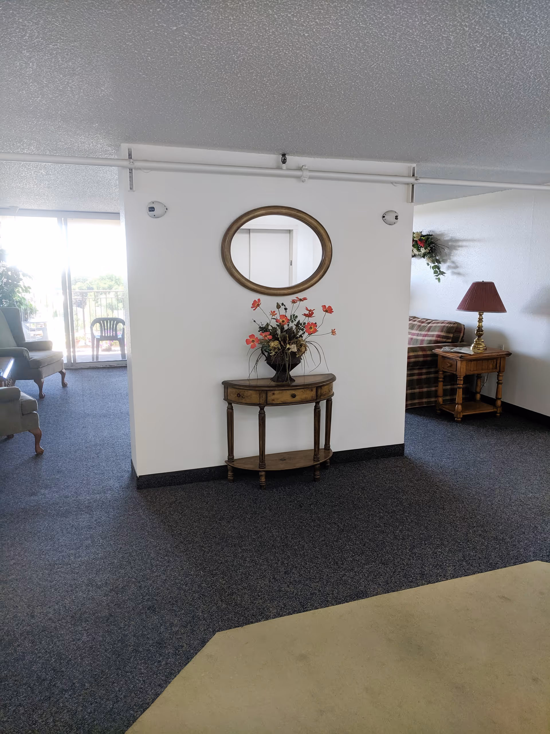 A white partition with a round mirror and a small wooden console table holding a floral arrangement, with seating and a lamp visible in the adjoining living area.