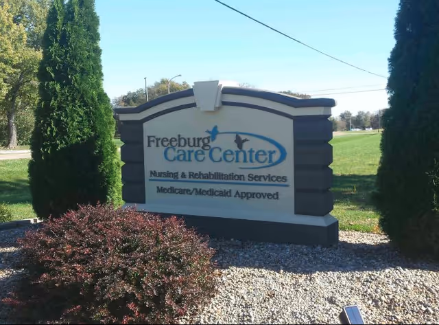 A landscaped roadside sign reading "Freeburg Care Center Nursing & Rehabilitation Services Medicare/Medicaid Approved" on a grassy lawn with trees.
