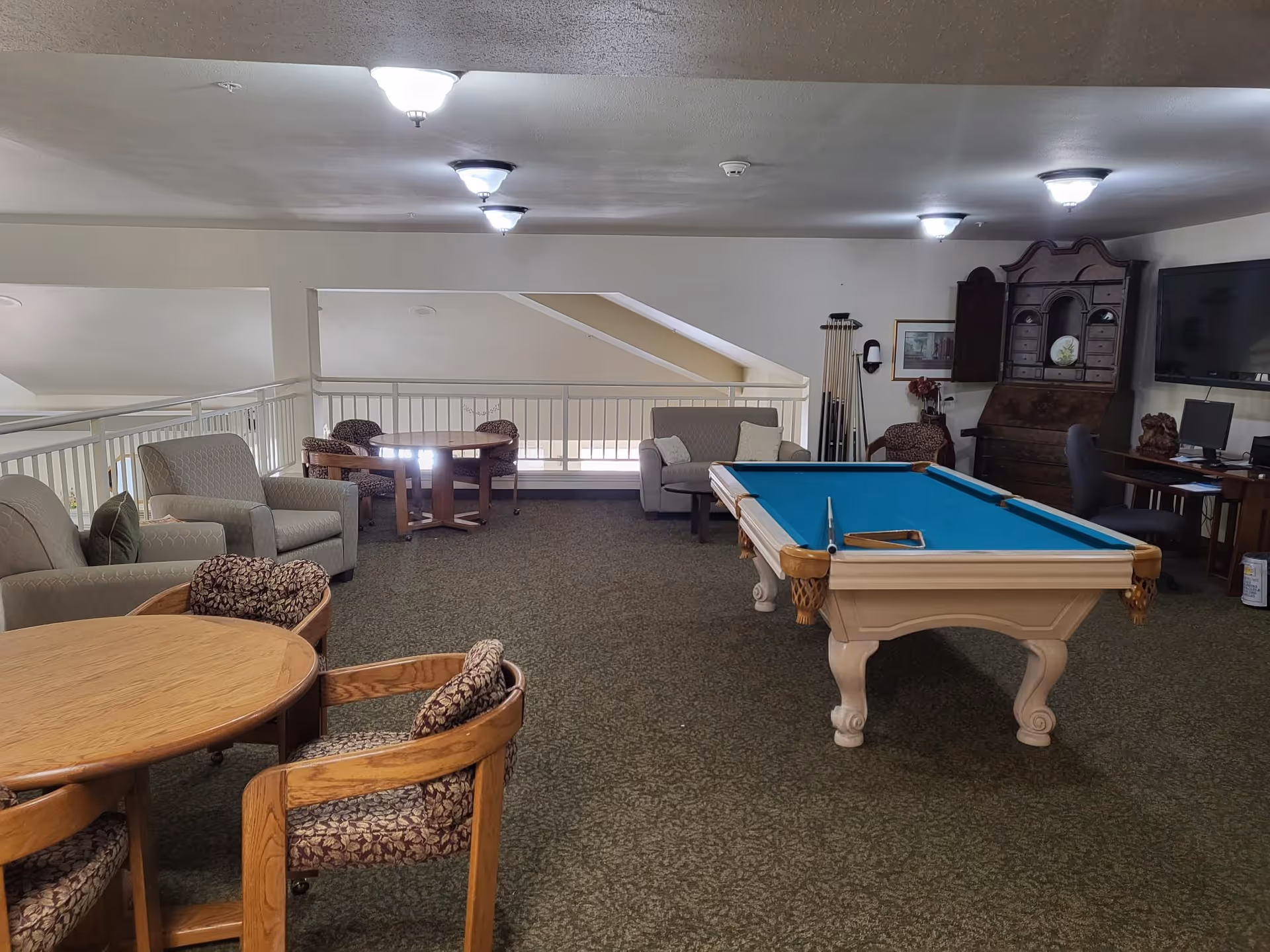 A recreational room with a pool table in the center, surrounded by various seating options including armchairs, cushioned chairs, and a small sofa. There are two round wooden tables with chairs, a wooden cabinet, a wall-mounted TV, and a computer desk with a chair. The room has carpeted flooring and ceiling lights.