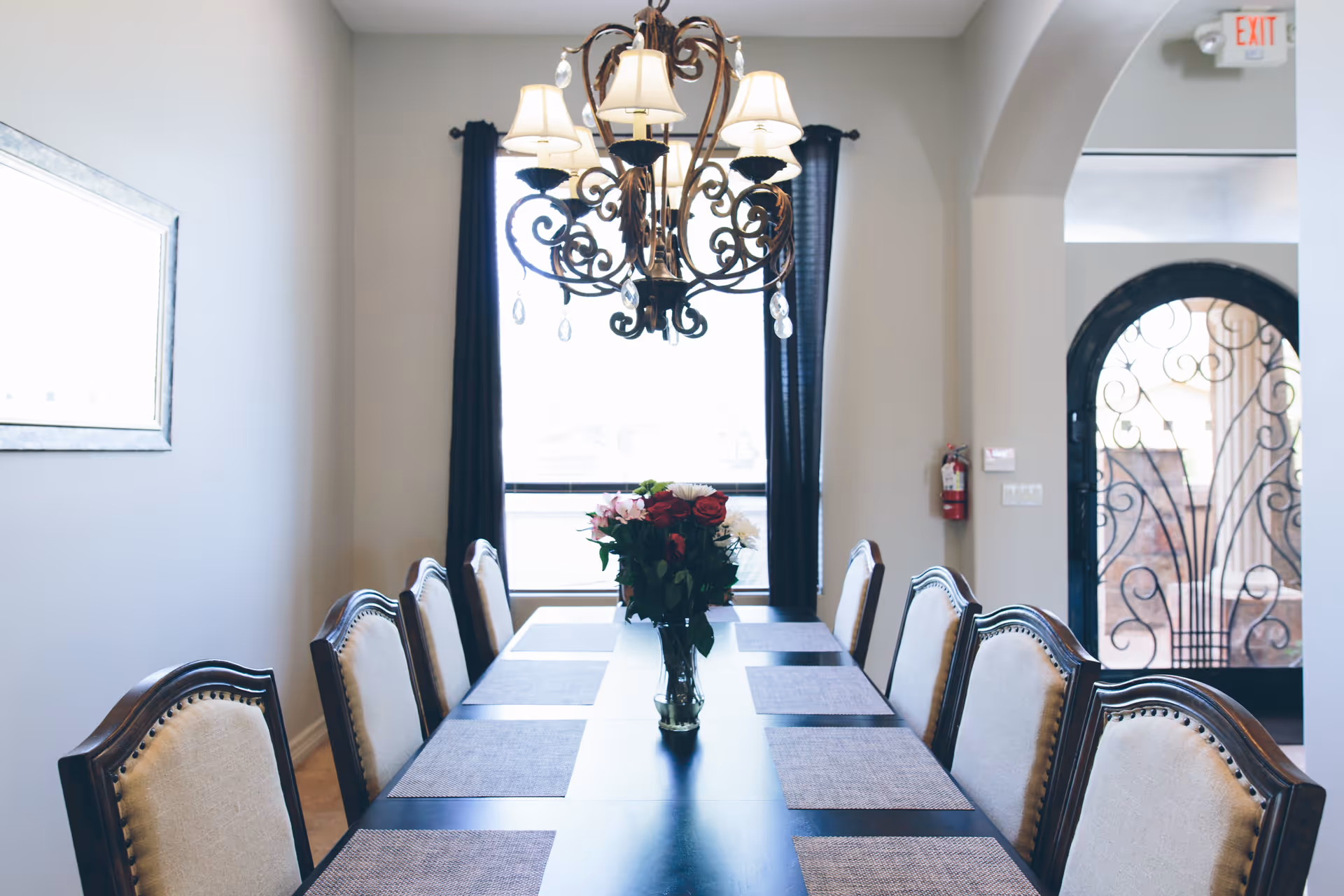 A dining room with a long dark wooden table set with placemats and a vase of flowers in the center. There are eight upholstered chairs around the table. A decorative chandelier hangs above the table, and a window with dark curtains is in the background. An arched doorway with a wrought iron gate is visible to the right.