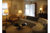 Cozy living room with a curved tufted sofa, armchairs, a glass coffee table, table lamps, and an open door letting in daylight.