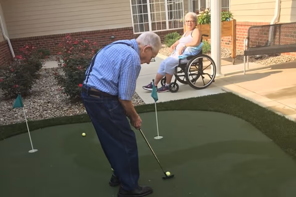 A man putting on a small outdoor putting green while a woman in a wheelchair watches in a courtyard.