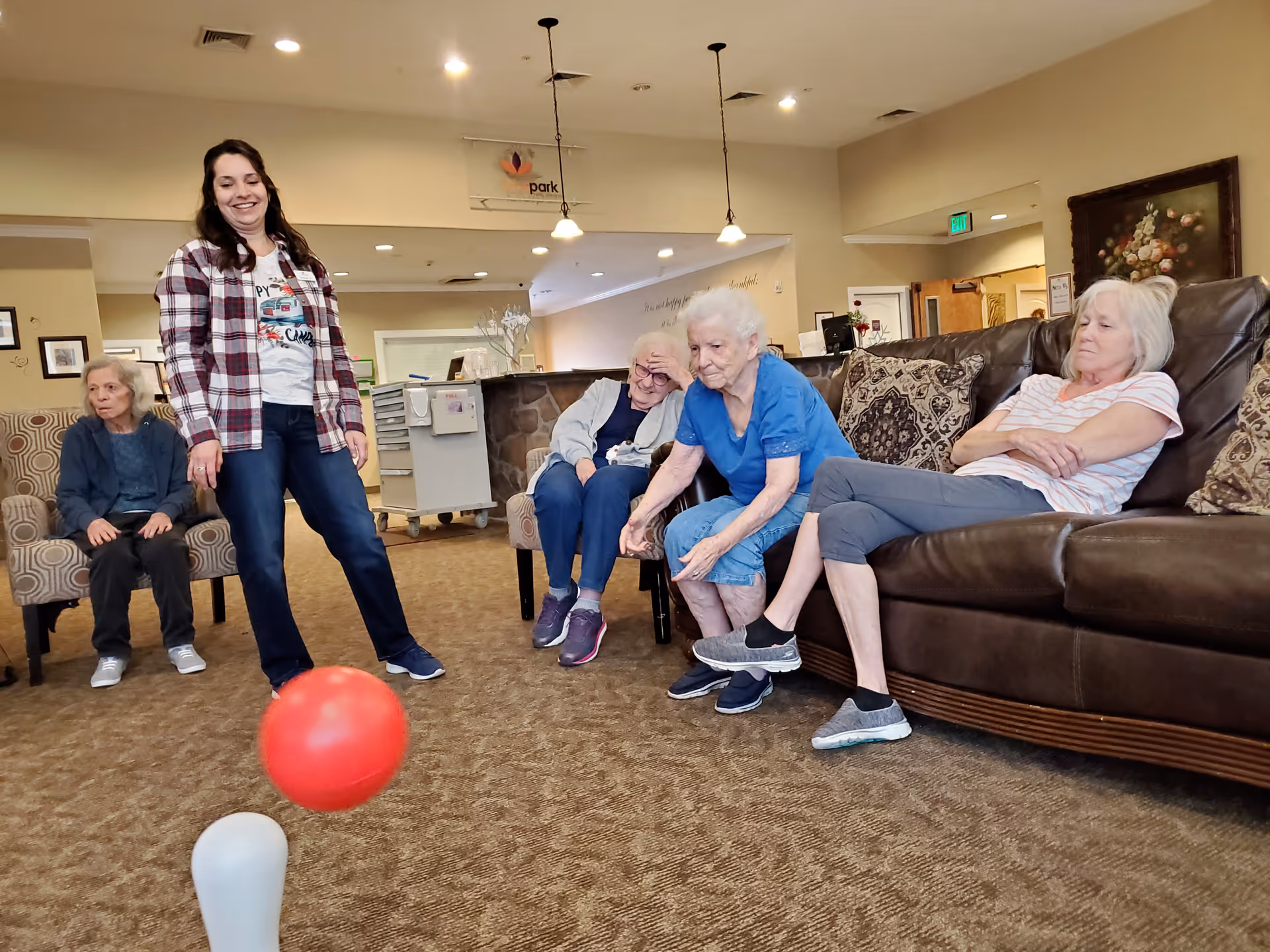 A group of elderly women and a younger woman are in a common area of a senior living facility. One elderly woman is sitting on a couch, another is seated on a chair, and a third elderly woman is reaching out towards a red ball on the carpeted floor. The younger woman is standing and smiling, watching the activity. The room has beige walls, pendant lights, and comfortable seating with patterned pillows.