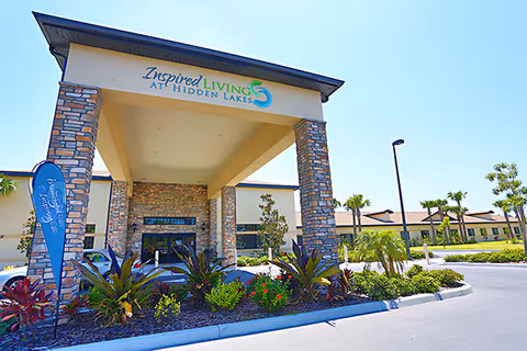 Front exterior view of the Inspired Living at Hidden Lakes facility with a large covered entrance supported by stone pillars, surrounded by landscaped plants and a clear blue sky.
