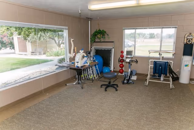 A rehabilitation room with exercise equipment including a stationary bike, weights, therapy balls, and a spine model. The room has large windows showing an outdoor view with greenery and a pathway.