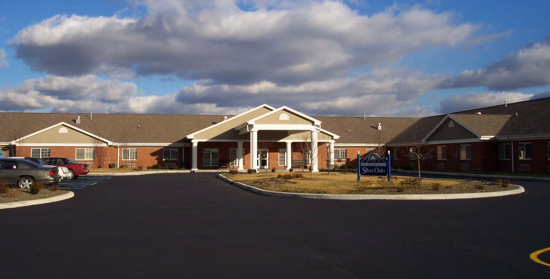 Exterior view of Silver Oaks Health Campus, a single-story brick building with a covered entrance supported by white columns, a circular driveway, and a sign displaying the facility name. Several cars are parked on the left side under a partly cloudy sky.