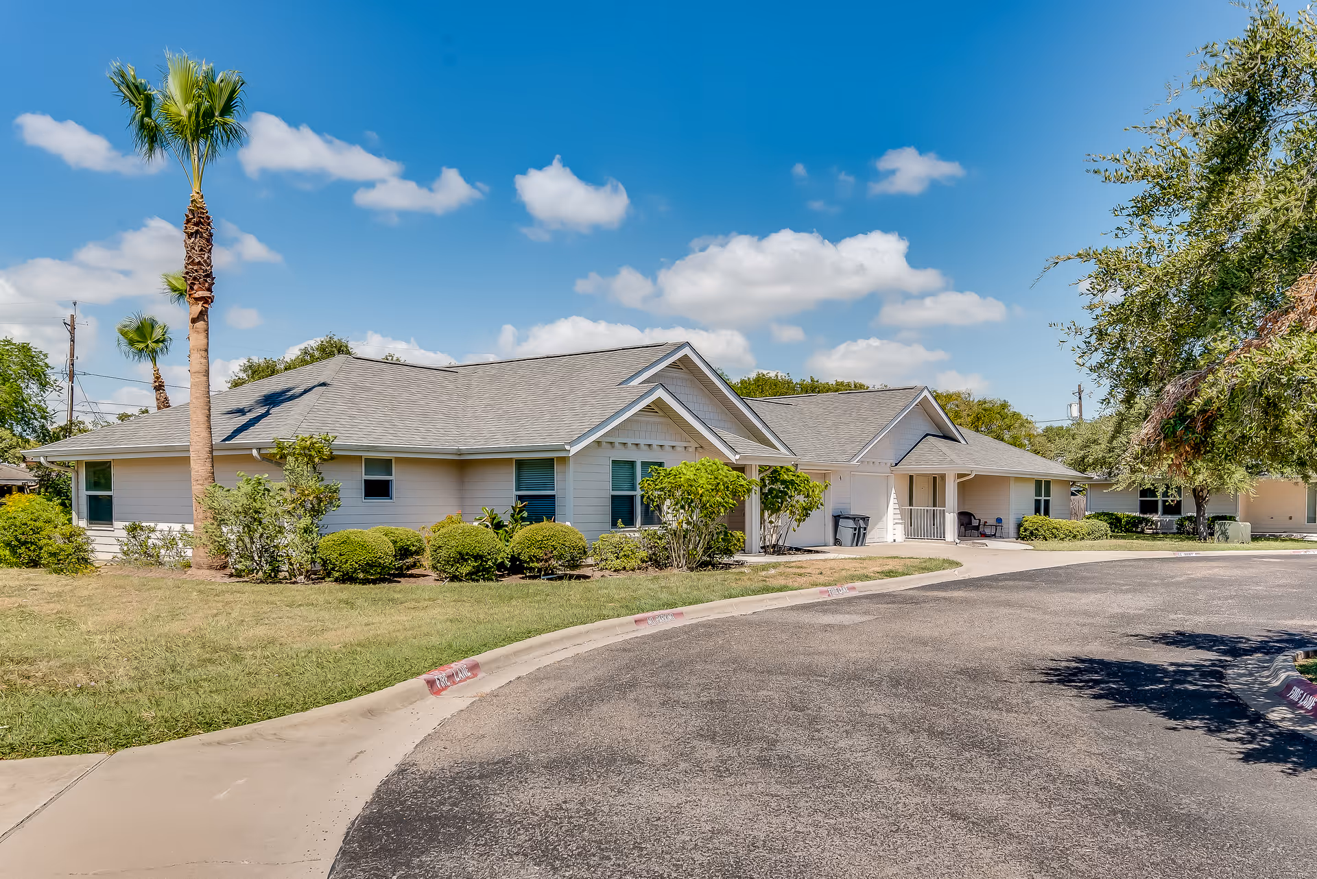Exterior view of a single-story senior living facility building with a gray roof, beige siding, and surrounded by green bushes and palm trees under a blue sky with scattered clouds.