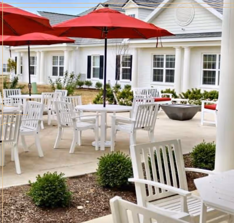 Outdoor patio area with white tables and chairs, red umbrellas providing shade, surrounded by small green bushes and a white building with black shutters in the background.