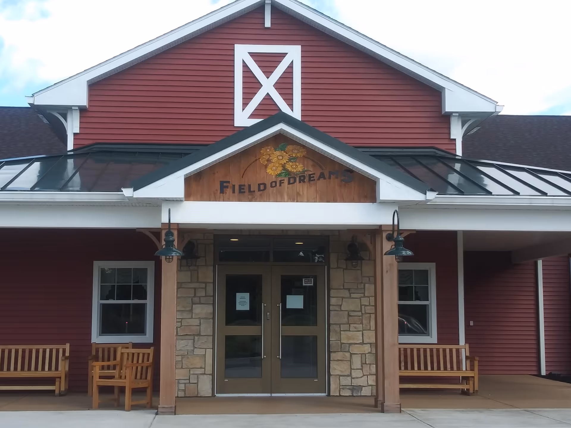 Front exterior view of a red building with white trim and a stone entrance. Above the double glass doors is a wooden sign with sunflowers and the text 'Field of Dreams'. There are two wooden benches on either side of the entrance and two green hanging lamps.