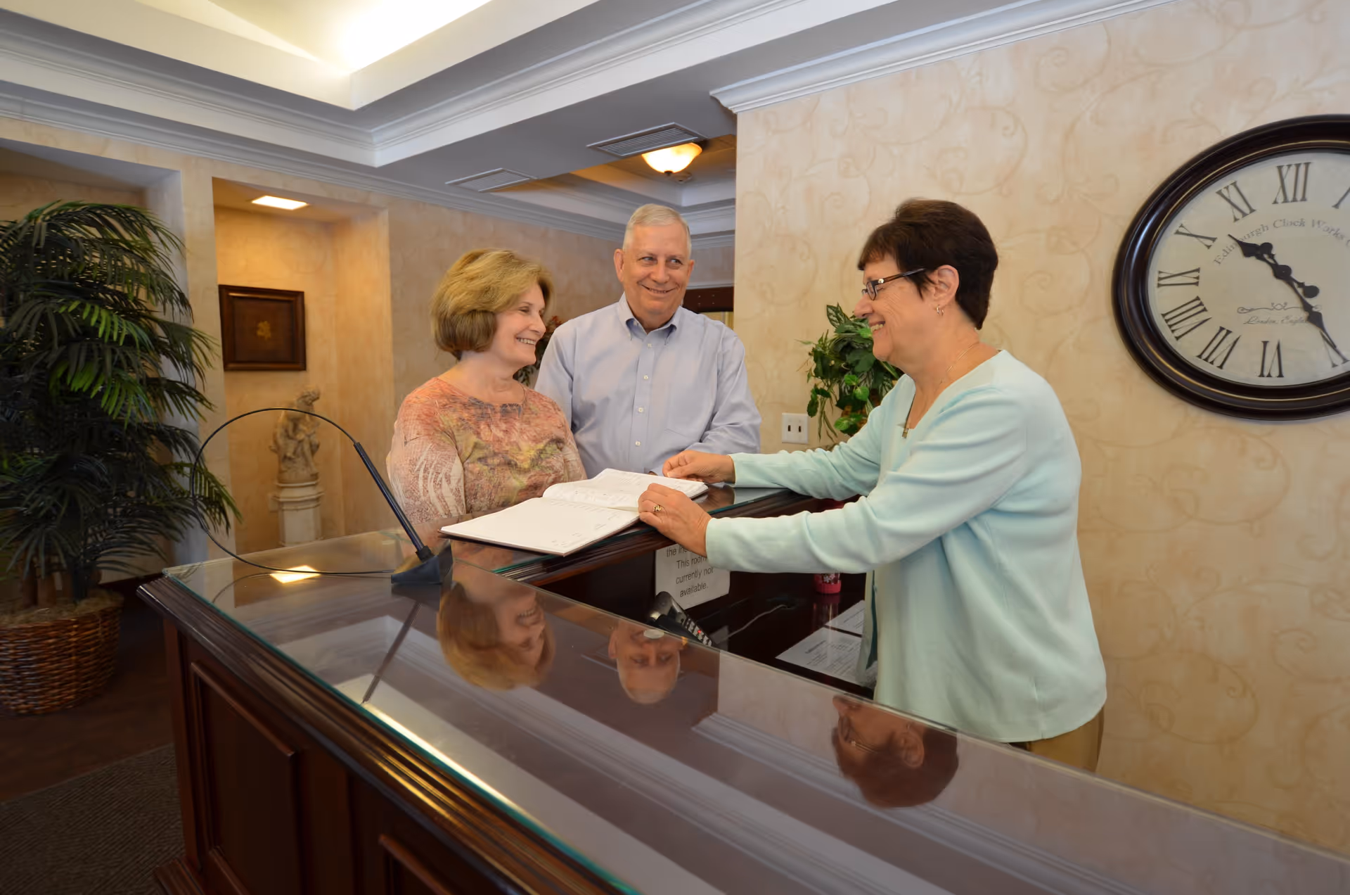 A woman behind a reception desk is assisting an elderly couple who are looking at an open book on the counter. The setting appears to be a welcoming indoor area with a large clock on the wall and a potted plant nearby.