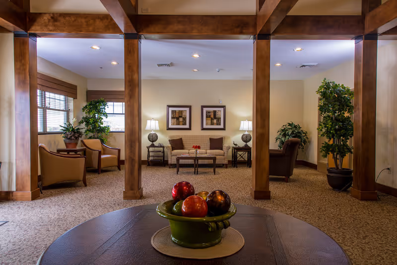 A cozy lounge with armchairs, a sofa, table lamps, wooden columns, and a decorative bowl of faux fruit on a table in the foreground.