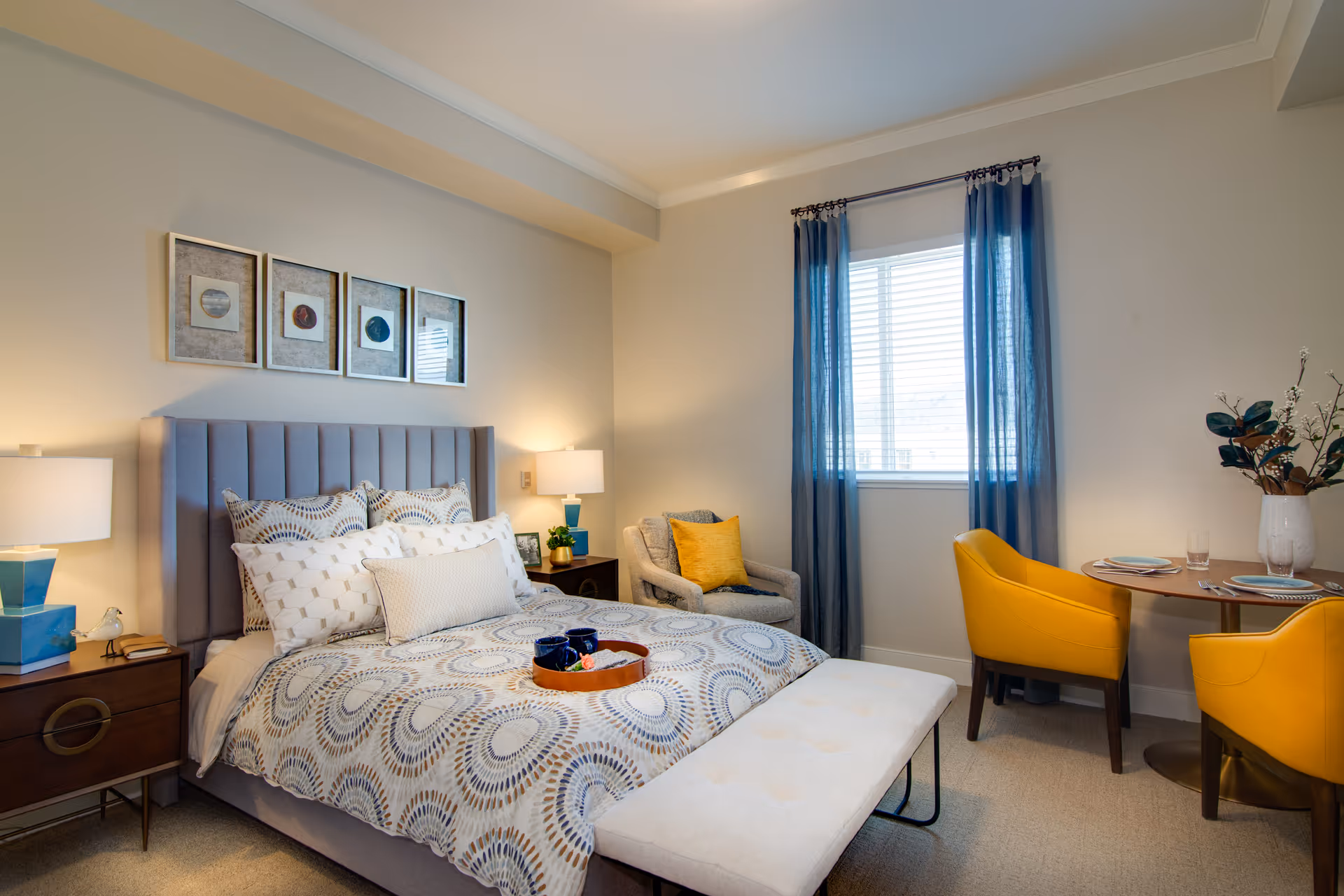 A cozy bedroom featuring a bed with a patterned comforter and multiple pillows, flanked by two nightstands each with a lamp. Above the bed are four framed artworks. A cushioned bench sits at the foot of the bed. To the right, there is a window with blue curtains, a small round dining table set with plates and glasses, and two yellow chairs. A comfortable armchair with a yellow pillow is positioned near the window. The room has neutral walls and carpeted flooring.