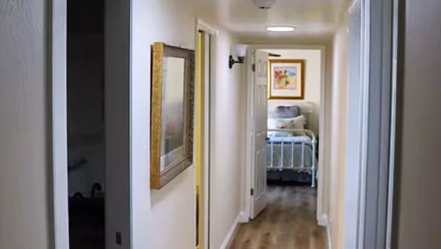 View down a narrow hallway with light-colored walls and wooden flooring leading to a bedroom with a bed, pillows, and a framed picture on the wall above the bed. There is a mirror on the left wall of the hallway and doors on both sides.