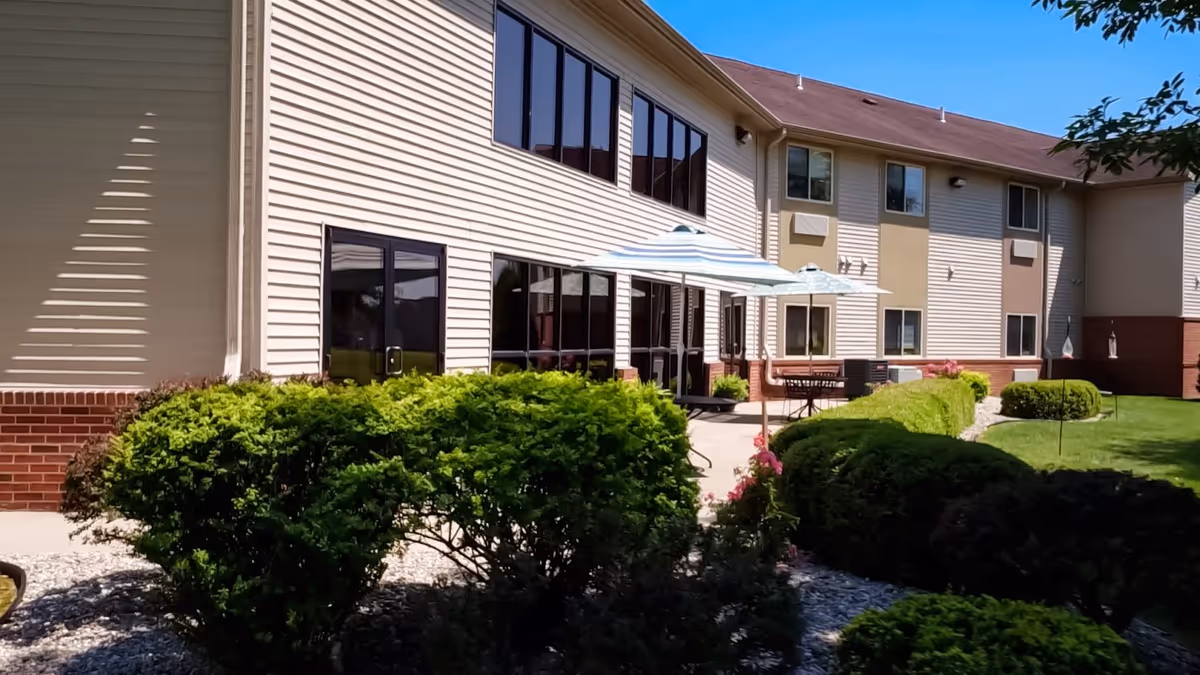 Outdoor patio area of a senior living facility with beige siding and brick accents. The patio has tables with umbrellas and is surrounded by well-maintained green bushes and a lawn. The building has multiple windows and a two-story structure.