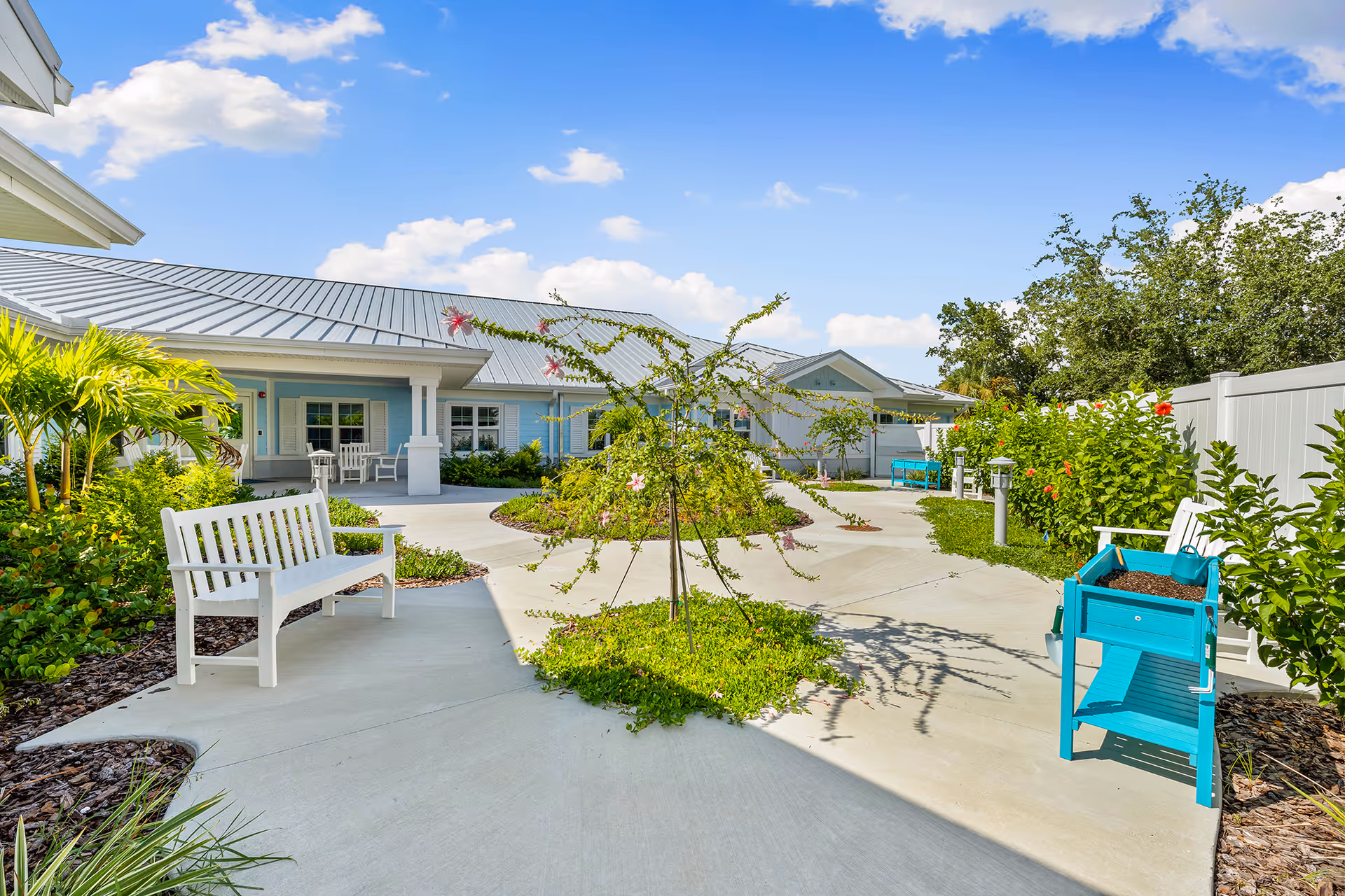Outdoor courtyard area at The Meridian at Punta Gorda Isles featuring a concrete pathway with white benches, green plants, small trees, and blue gardening planters under a bright blue sky with some clouds.