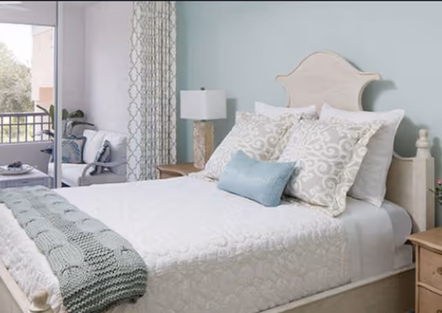 A neatly made bed with a white quilt, decorative pillows, and a light blue throw blanket in a bedroom. Next to the bed is a wooden nightstand with a lamp. In the background, there is a sliding glass door leading to a balcony with outdoor seating and a view of trees.