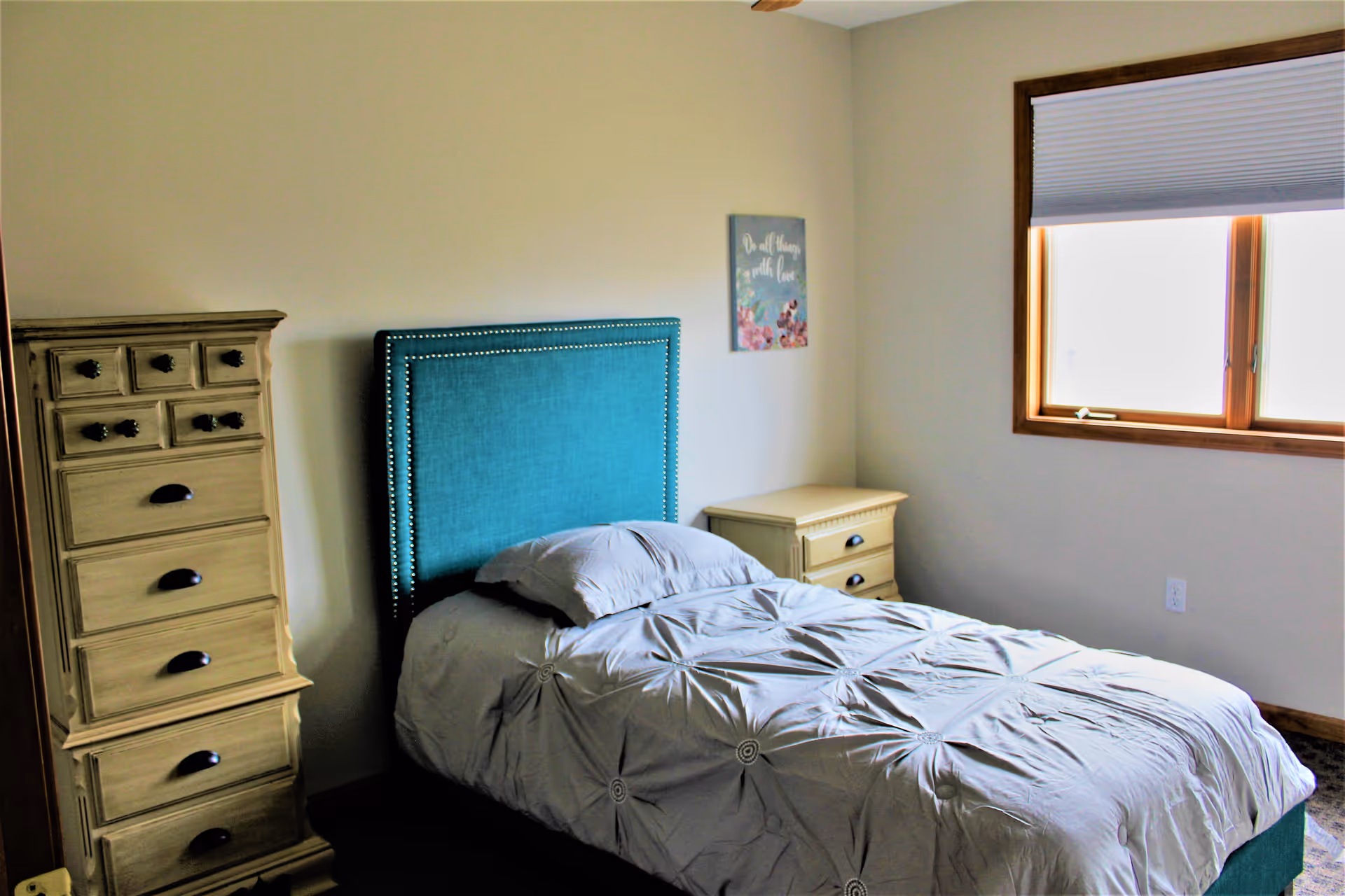 A bedroom with a single bed featuring a teal upholstered headboard and a light gray comforter. To the left of the bed is a tall wooden dresser with multiple drawers, and to the right is a small wooden nightstand with two drawers. A window with a closed blind is on the right wall, and a decorative wall art with floral design and the text 'Do all things with love' hangs above the nightstand.
