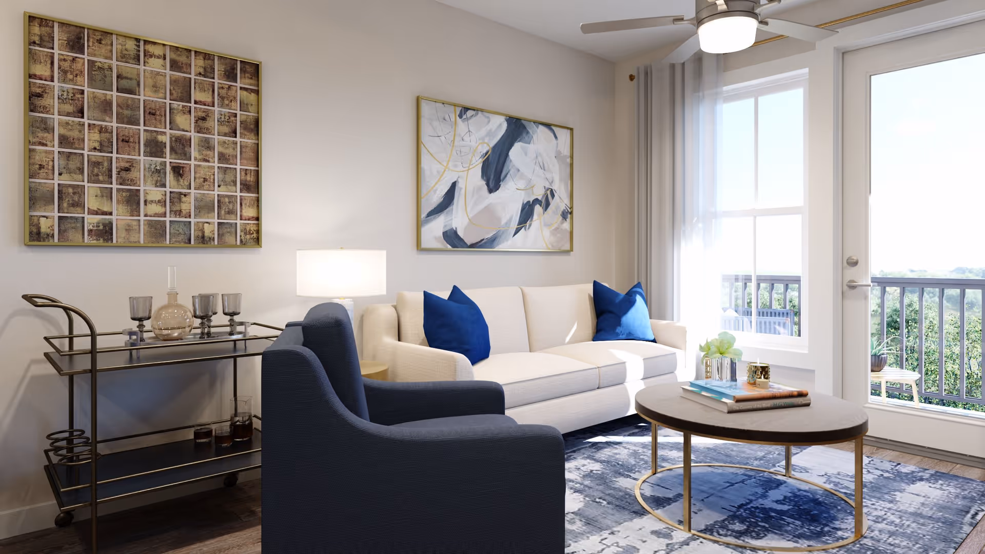 A bright living room with a beige sofa adorned with two blue pillows, a dark blue armchair, a round coffee table with books and a small plant, a bar cart with glassware and a lamp, two abstract paintings on the wall, a ceiling fan with a light, and a large window and glass door leading to an outdoor balcony with greenery visible outside.
