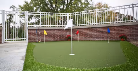 A small outdoor putting green with four colored flags (white, yellow, red, and blue) surrounded by a white metal fence and a brick wall, with trees visible in the background.