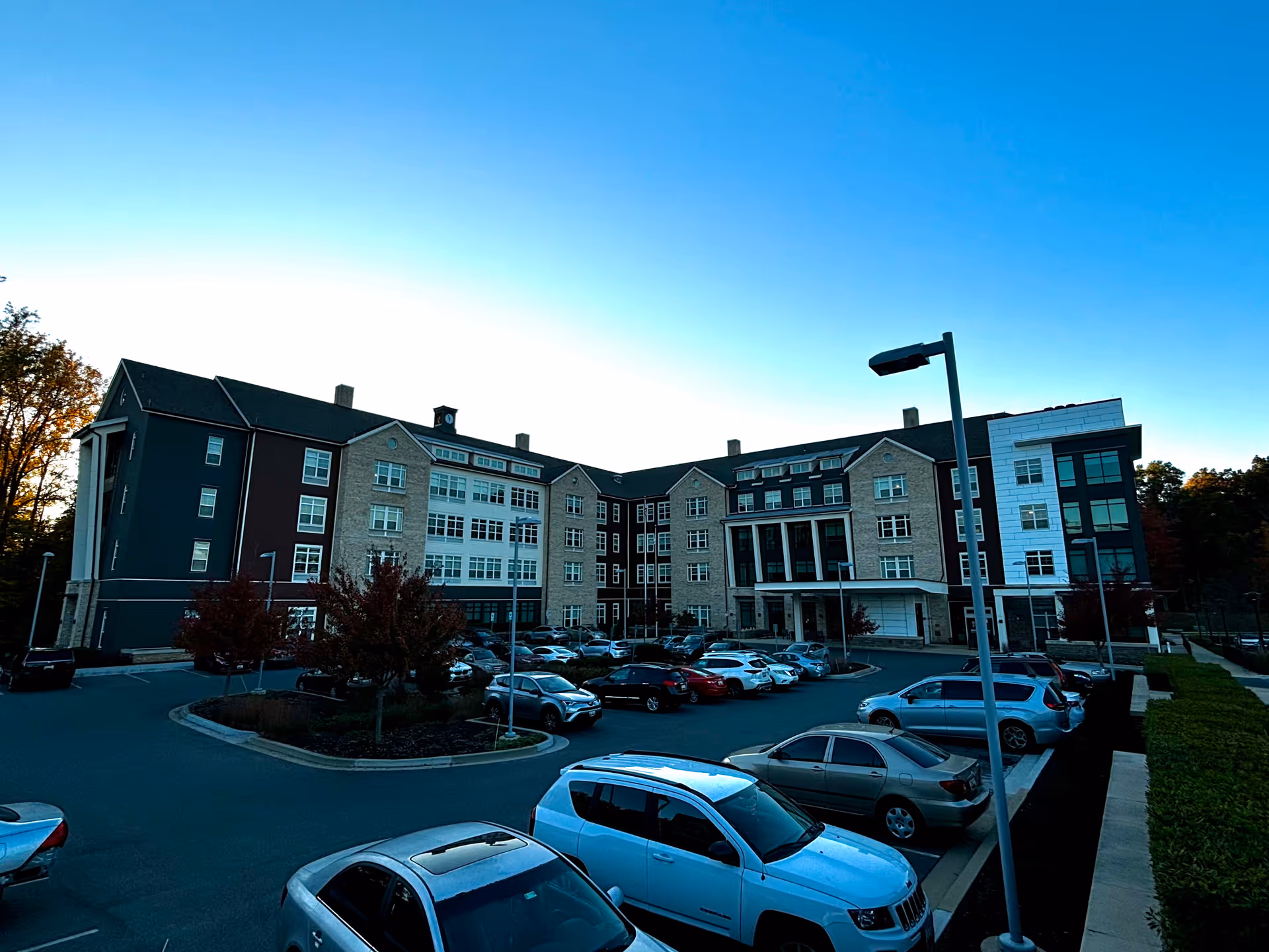 Exterior view of a large multi-story senior living facility building named Tribute at Black Hill during dusk, with a parking lot filled with cars in the foreground and trees in the background under a clear sky.