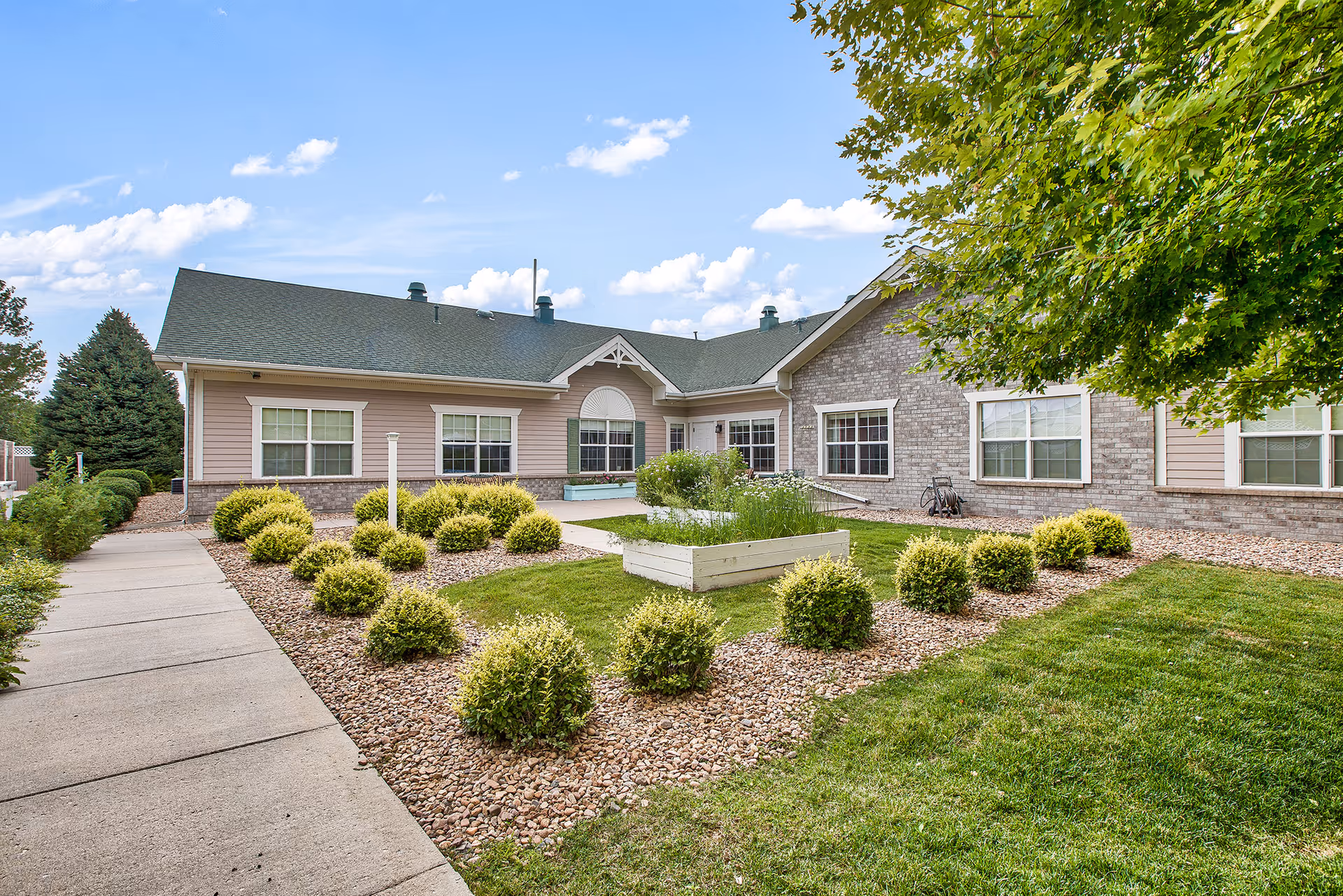Exterior view of a single-story senior living facility building with a green roof, beige and brick walls, surrounded by a landscaped garden with bushes, a raised planter box, and a concrete walkway under a partly cloudy sky.