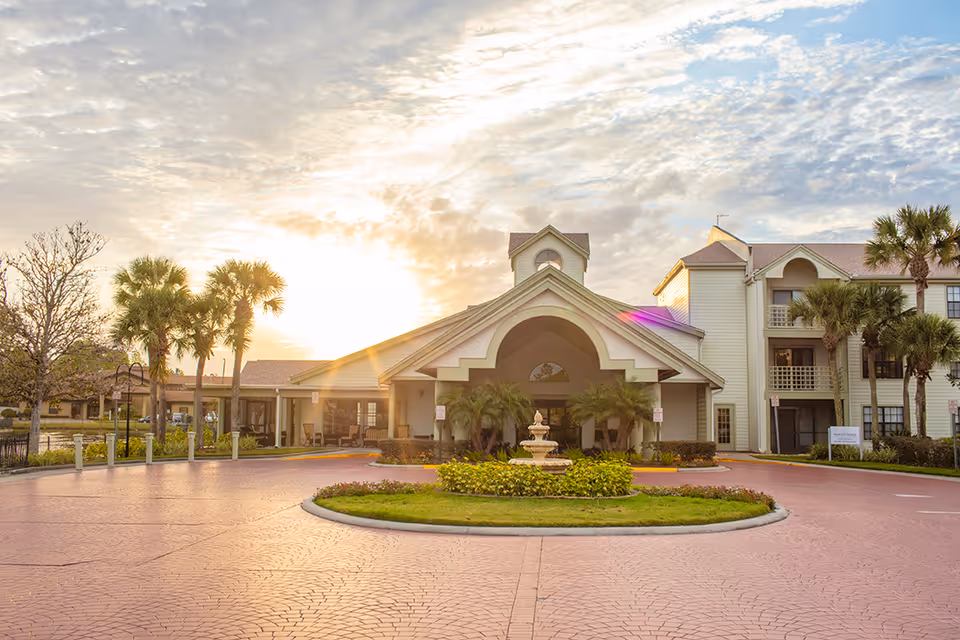 Front exterior view of Cascade Heights facility with a circular driveway, landscaped greenery, palm trees, and a fountain in the center. The building has a large covered entrance with an arch and multiple windows, under a partly cloudy sky with the sun setting or rising.