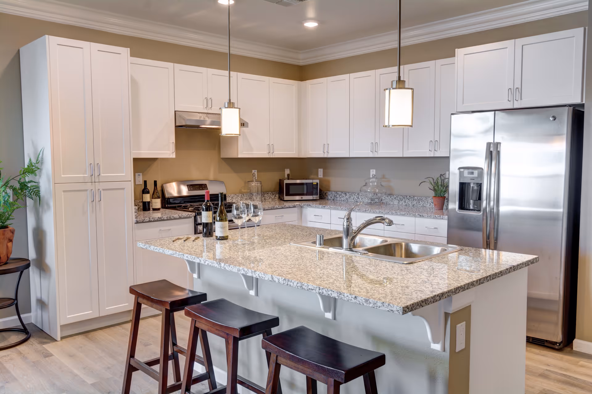 Modern kitchen with white cabinets, granite countertops, and stainless steel appliances including a refrigerator and stove. There is a kitchen island with a sink, three wooden bar stools, and two pendant lights hanging above. Wine bottles and glasses are placed on the island, and a small plant is on the counter near the refrigerator.