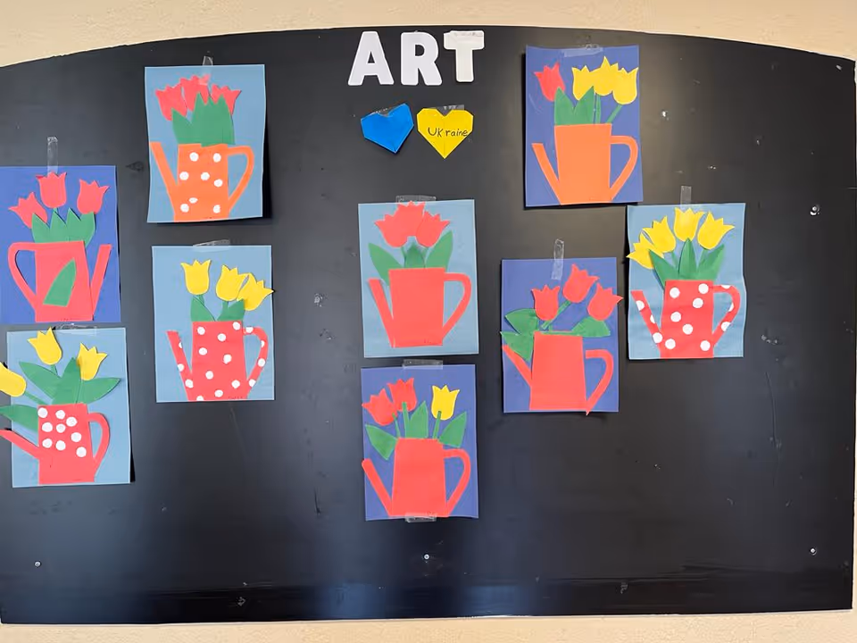 A black bulletin board with the word 'ART' at the top displaying colorful paper cut-out artwork of watering cans with tulips in red, yellow, and green. Two paper hearts, one blue and one yellow with the word 'Ukraine' written on it, are also pinned on the board.