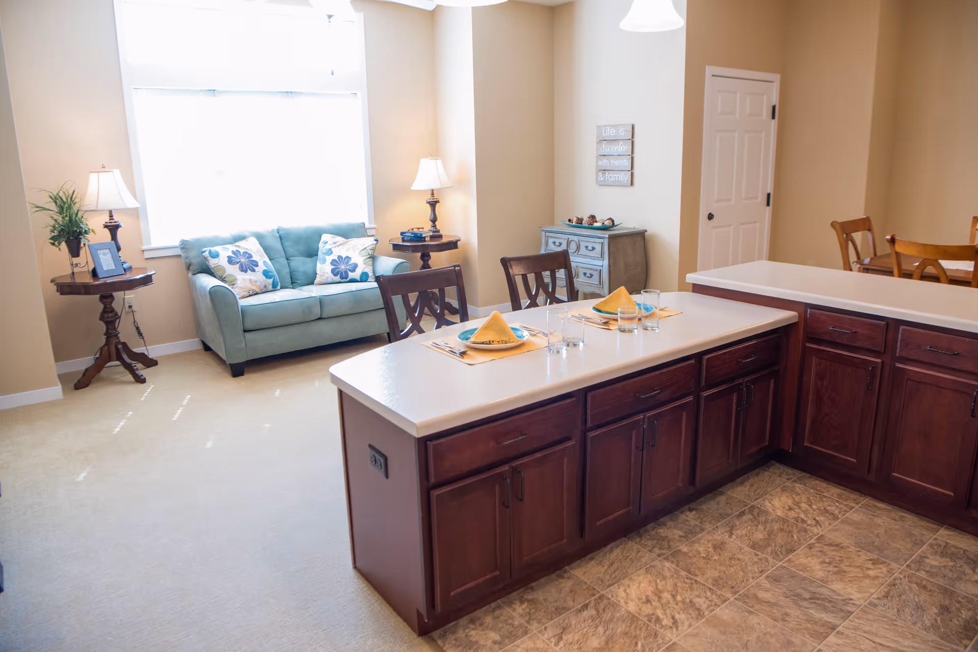 A bright and cozy living area with a light blue loveseat adorned with floral pillows, two side tables with lamps and a plant, and a kitchen island with place settings for two. The room has beige walls, a tiled floor near the kitchen, and carpet in the living area. A small chest of drawers and a door are visible in the background.